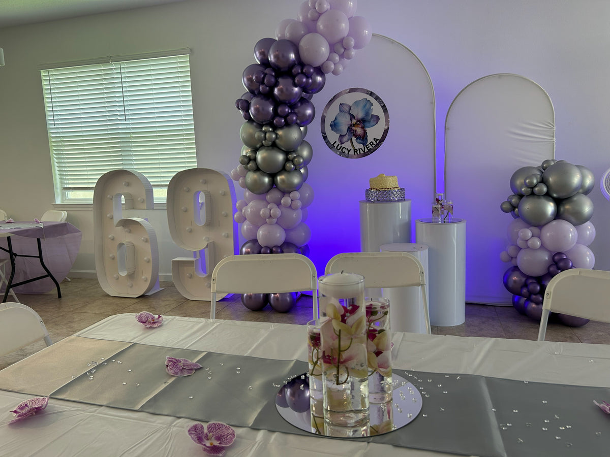 Lavender and silver birthday decoration with two white arch backdrops round personalized sticker cylinder pedestals and purple accent lights.