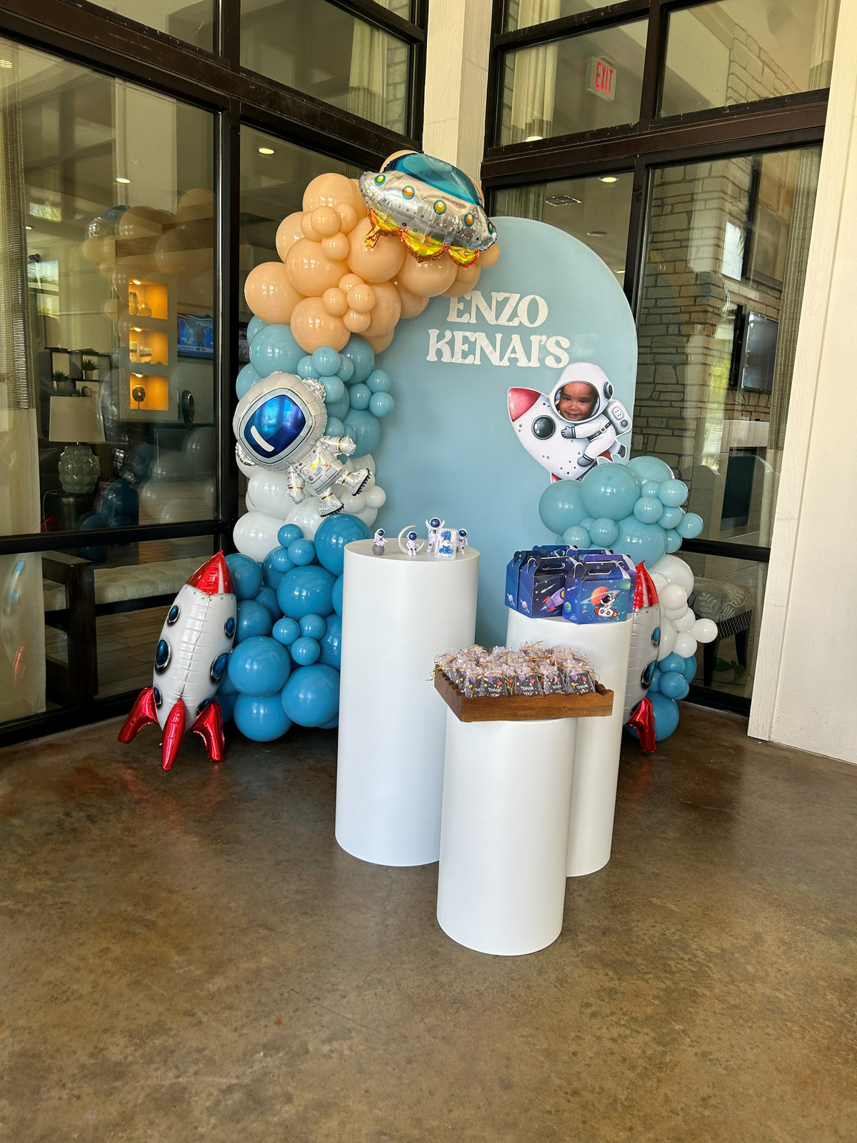 Blue space birthday decoration with rockets astronaut balloons arch backdrop and white cylinder pedestals.