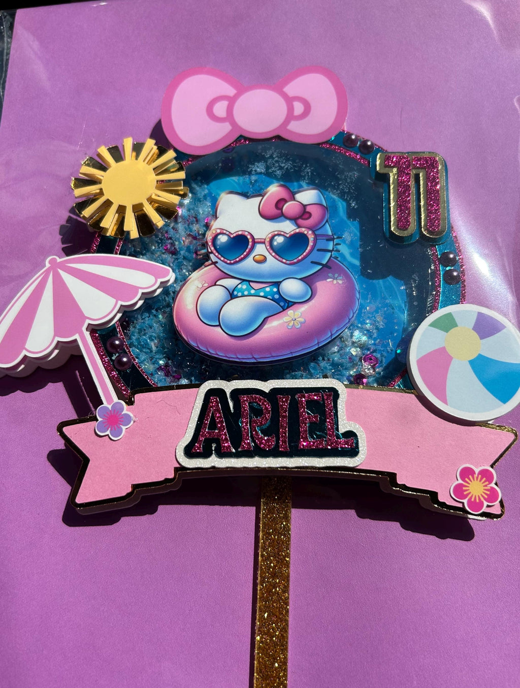 Close view of a pink and blue kitty pool party topper with Ariel name banner and age eleven against a purple background.