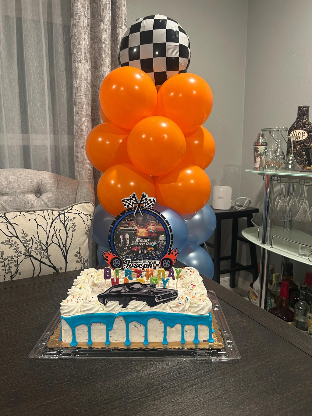 Racing birthday cake with personalized topper set beneath an orange and gray balloon column and checkered balloon.