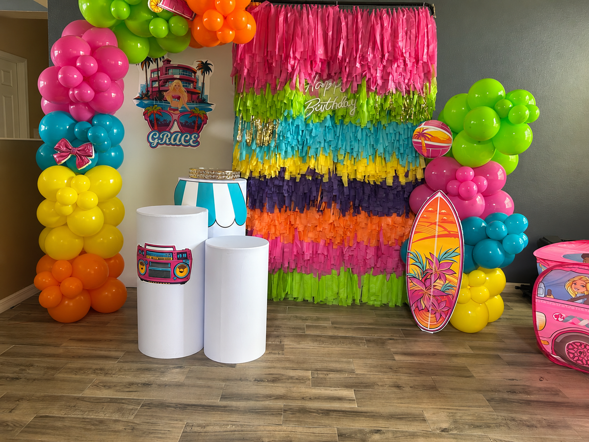 Tropical balloon decoration with color block balloon columns, white backdrop, multicolor fringe, three white cylinder pedestals and Happy Birthday sign.